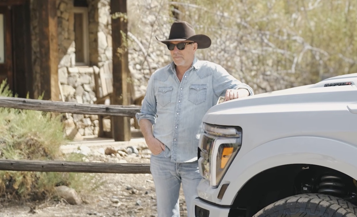 Still from the Lifted Trucks video: Kevin Costner in a cowboy hat leaning on a white pickup truck outside a stone cabin