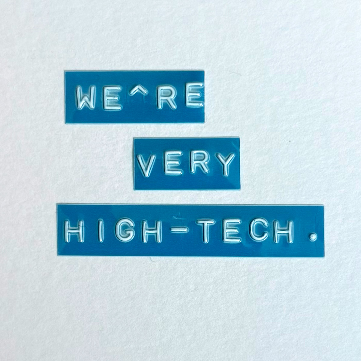 Blue embossed label-maker tape on white paper spelling: we’re very high-tech.