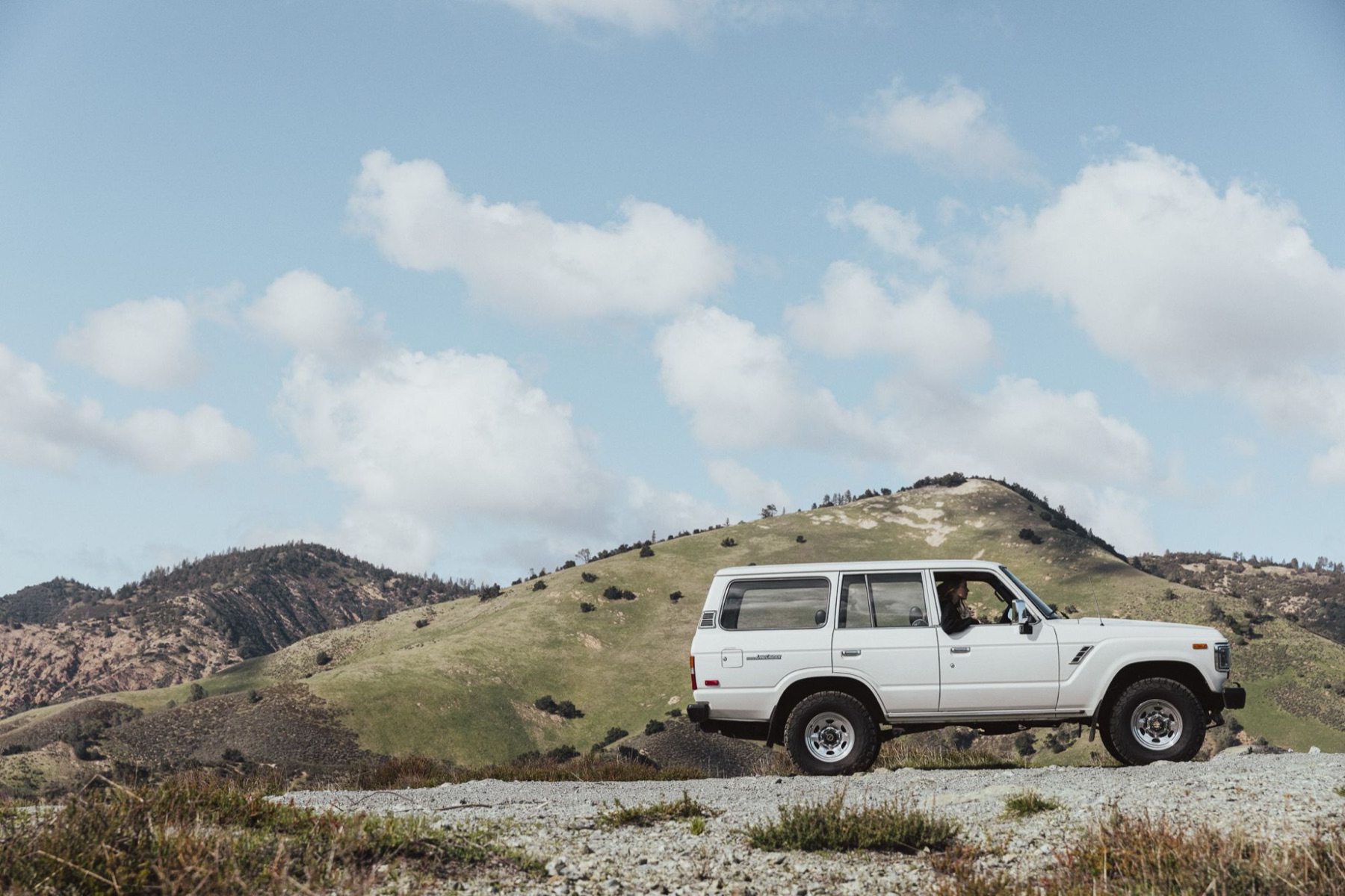 White vintage Toyota Land Cruiser on a gravel road below green hills